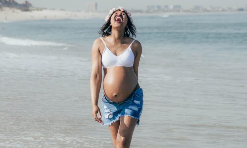 Femme enceinte heureuse marchant sur la plage, pieds dans l'eau. Photographe : Leo Moko.